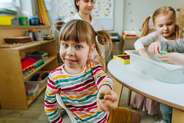 Learning play activities in kindergarten or daycare centre. Preschool students looking for wooden beads in a container with kinetic sand and sorting it by color. Education concept. Selective focus. - Foto, imagen