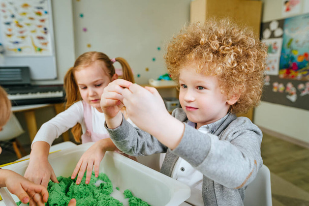 Learning play activities in kindergarten or daycare centre. Preschool students looking for wooden beads in a container with kinetic sand and sorting it by color. Education concept. Selective focus. - Photo, Image