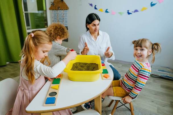 Preschool students looking for wooden geometric forms in a container with raw mung beans and sorting it. Learning play activities in kindergarten or daycare centre. Education concept. Selective focus. - 写真・画像