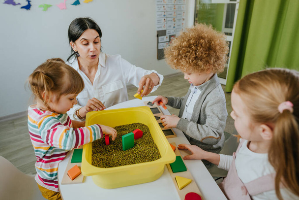 Preschool students looking for wooden geometric forms in a container with raw mung beans and sorting it. Learning play activities in kindergarten or daycare centre. Education concept. Selective focus. - Foto, Bild