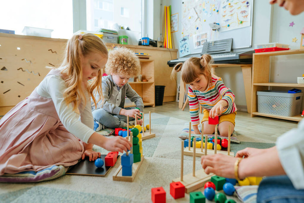 Preschool students sorting wooden geometric toy balls and cubes sittings on the floor. Learning toys in kindergarten or daycare centre. Education concept. Selective focus. - 写真・画像