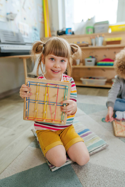 Adorable girl playing with geoboard wrapping rubber bands. Kind of mathematical manipulative made from wooden board. Learning toys in kindergarten. Education concept. Selective focus. - Valokuva, kuva
