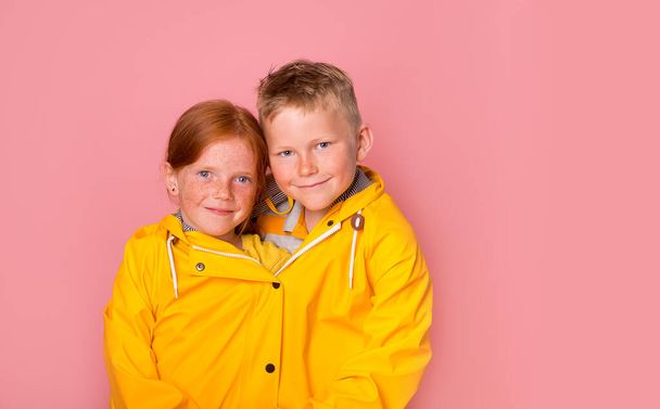 Funny children girl and boy in one yellow rain coat on color pink background. Sale discount. Two in one. Portrait of happy ginger freckled kids. School stylish child. Twins. Fashion. advertising - Φωτογραφία, εικόνα