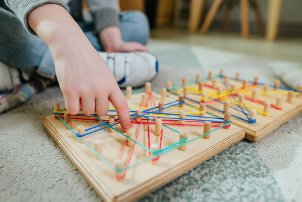 Close up of boy playing with geoboard wrapping rubber bands. Kind of mathematical manipulative made from wooden board. Selective focus. - Photo, image