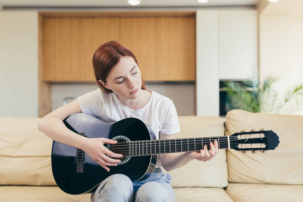 Young beautiful red-haired woman sitting at home on the couch and playing acoustic guitar, smiling and rejoicing - Фото, изображение