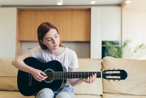 Молодая красивая рыжая женщина сидит дома на диване и играет на акустической гитаре, улыбаясь и радуясь - Фото, изображение