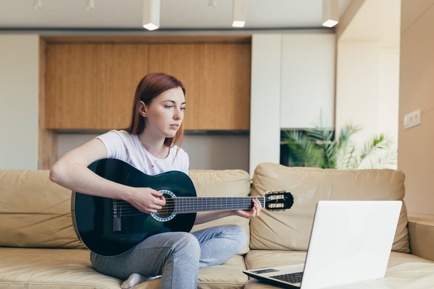 young red haired girl learns to play the guitar with the help of video tutorial lessons. female woman home at leisure sitting on sofa with laptop studying online musical instrument. Hobbies distance - 写真・画像