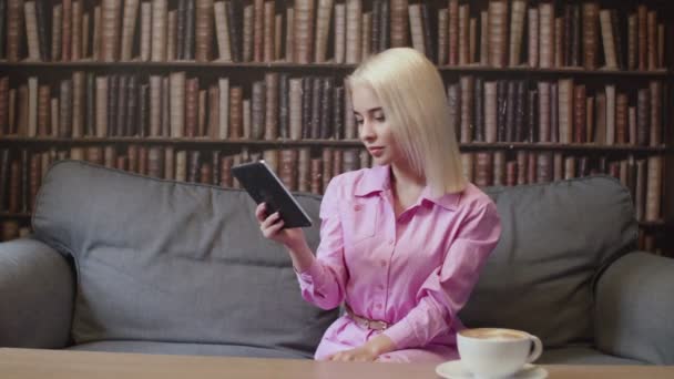 A menina está sentada na biblioteca e lendo um e-book - Filmagem, Vídeo