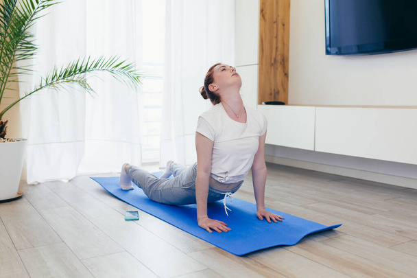 jovem mulher atraente está praticando ioga on-line usando um telefone celular ou aplicativo de smartphone. Manhã de yogi feminino em roupas casuais em casa na sala de estar, posição de lótus treino medita em um tapete - Foto, Imagem