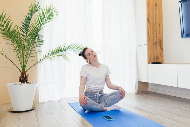 young attractive woman is practicing yoga online using a mobile phone or smartphone app. Female yogi morning in casual clothes at home in living room, workout lotus position meditates on a mat - Valokuva, kuva