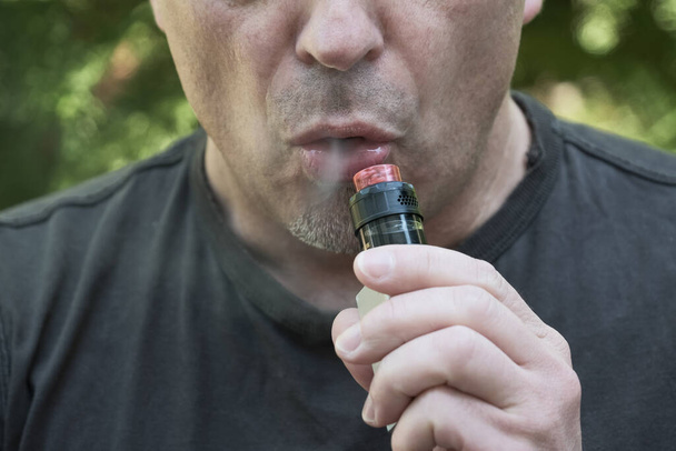 Лицо человека, курящего электронную сигарету. Мужчина держит в руках вапельное устройство и выдыхает дым. Выпаривание, вдыхание жидкости, популярная альтернатива курению. - Фото, изображение