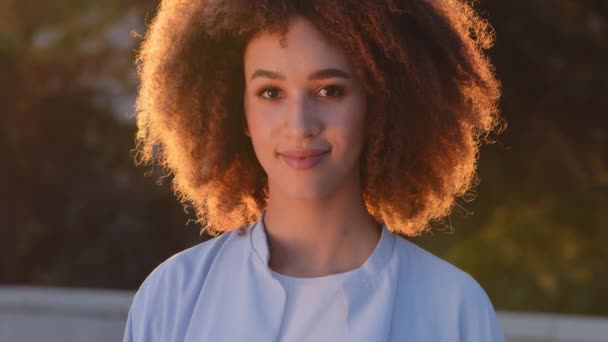 Nő szabadban portré gyönyörű fiatal csábító etnikai afro amerikai hölgy afro mosolygós lány göndör modell nő pózol az utcán napfényben napsugarak naplemente nézi kamera mosolyog aranyos barátságos - Felvétel, videó