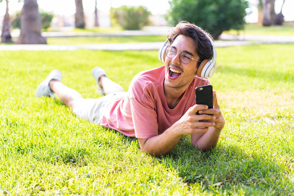 Young caucasian man at outdoors in a park listening music with the mobile and happy - Photo, image