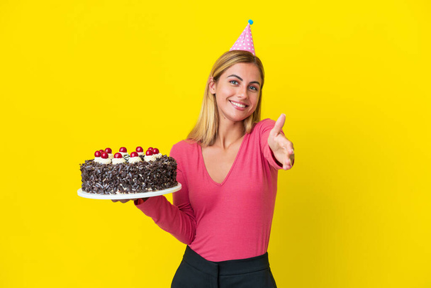 Sarışın Uruguaylı kız sarı arka planda izole edilmiş doğum günü pastası tutuyor ve iyi bir anlaşma için el sıkışıyor. - Fotoğraf, Görsel