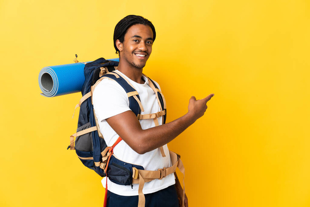 Joven montañero con trenzas con una gran mochila aislada sobre fondo amarillo apuntando hacia atrás - Foto, imagen