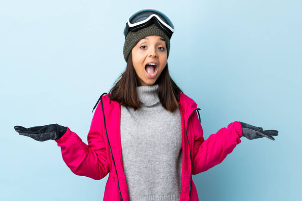Gemischte Rennläuferin mit Snowboardbrille vor isoliertem blauen Hintergrund mit schockiertem Gesichtsausdruck. - Foto, Bild