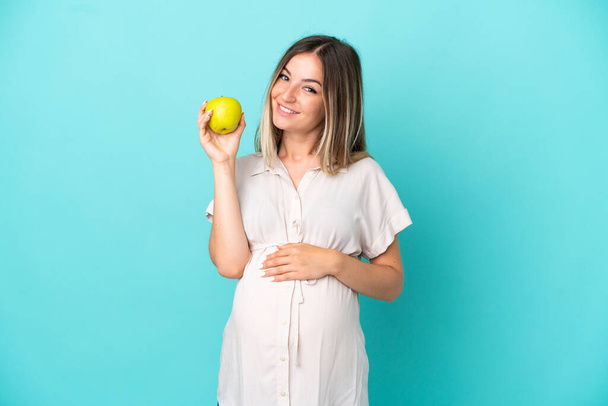 Junge rumänische Frau isoliert auf blauem Hintergrund schwanger und hält einen Apfel - Foto, Bild