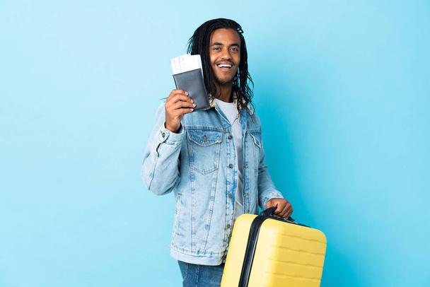 Young African American man with braids isolated on blue background in vacation with suitcase and passport - Photo, Image