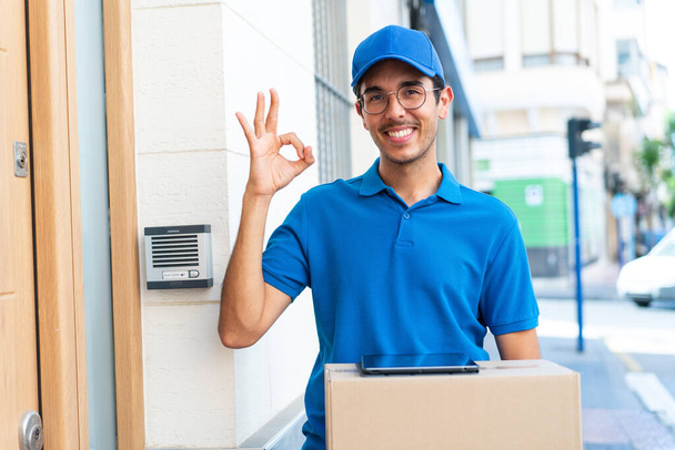 Молодой доставщик на открытом воздухе держит коробки и планшет и делает ОК знак - Фото, изображение