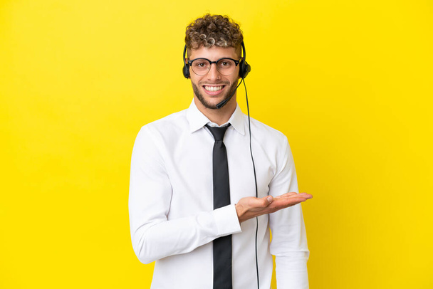 Telemarketer blonde man werken met een headset geïsoleerd op gele achtergrond presenteren een idee terwijl het kijken glimlachend in de richting van - Foto, afbeelding