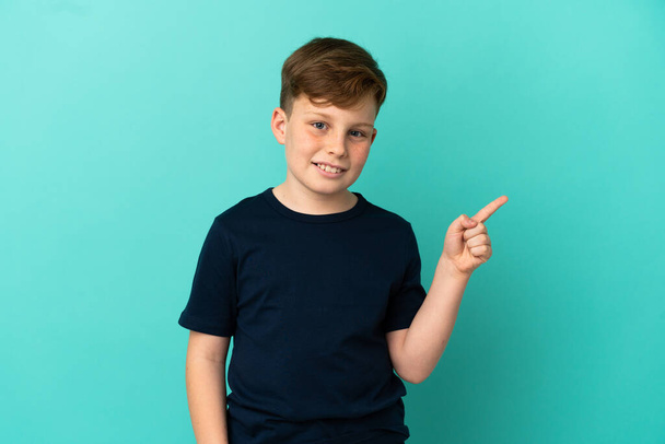 Little redhead boy isolated on blue background happy and pointing up - 写真・画像