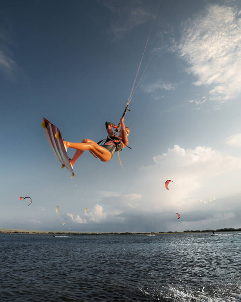 Professional athlete kitesurfer young caucasian woman doing a trick in the air against the backdrop of the sunset sky and clouds. Professional kitesurfing and kite culture training - Foto, afbeelding