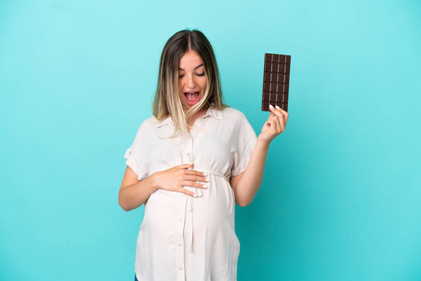 Young Romanian woman isolated on blue background pregnant and holding chocolate with surprised expression - Fotó, kép