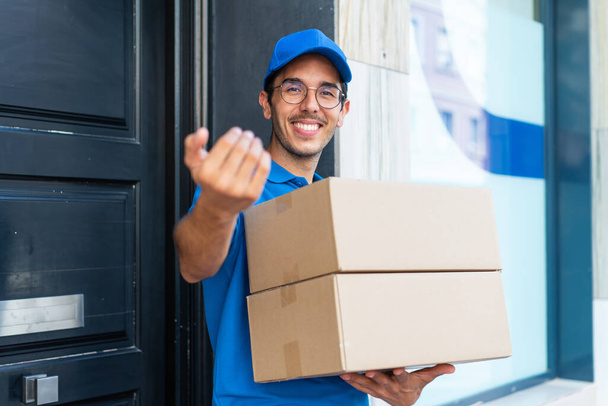 Молодой доставщик на открытом воздухе держит коробки со счастливым выражением лица и делает предстоящий жест - Фото, изображение