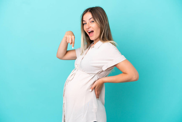 Young Romanian woman isolated on blue background pregnant and pointing to the belly - Foto, Imagen