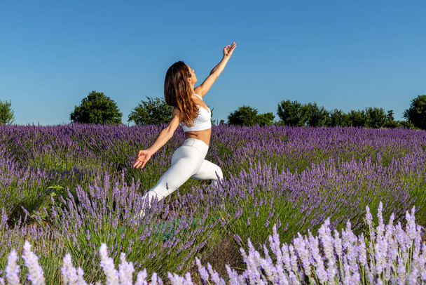 Guerriero posa yoga in un campo di lavanda in fiore. Una donna con lunghi capelli castani allunga la parte anteriore del corpo per rafforzare le gambe, il busto e la schiena. Senso di energia e stabilità. - Foto, immagini
