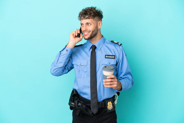 Young police blonde man isolated white on blue background holding coffee to take away and a mobile - Fotografie, Obrázek