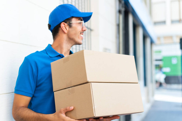 Young delivery man at outdoors holding boxes with happy expression looking side - Photo, Image