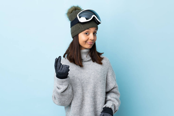 Змішана гоночна лижниця дівчина зі сноубордистськими окулярами над ізольованим синім тлом, що заробляє грошовий жест
. - Фото, зображення