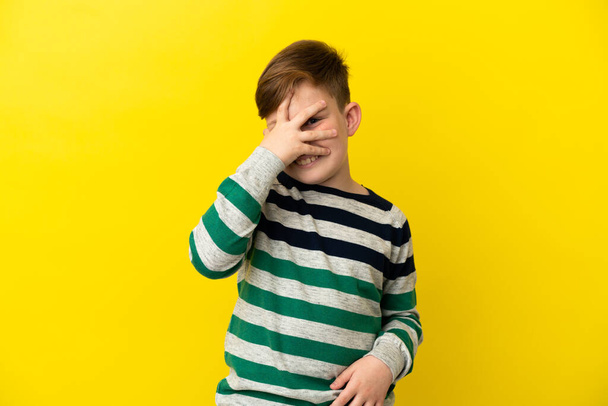 Little redhead boy isolated on yellow background covering eyes by hands and smiling - Photo, Image