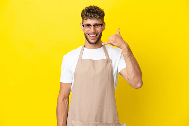 Restaurant waiter blonde man isolated on yellow background making phone gesture. Call me back sign - Photo, Image