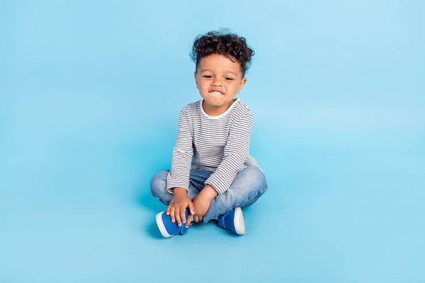 Portrait of attractive cheerful boy sitting on floor isolated over bright blue color background - Foto, afbeelding