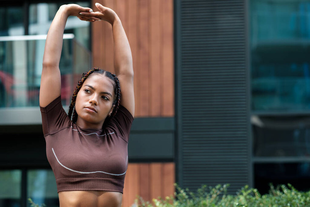 Fitness young woman wearing brown sportswear and stretching in urban environment. Mixed race, braid. - Foto, imagen