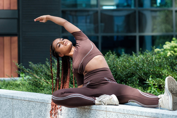 Fitness young woman wearing brown sportswear and stretching in urban environment. Mixed race, braid. - Photo, Image
