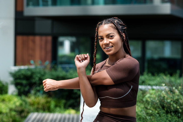 Mujer joven fitness con ropa deportiva marrón y estiramiento en el entorno urbano. Raza mixta, trenza. - Foto, Imagen