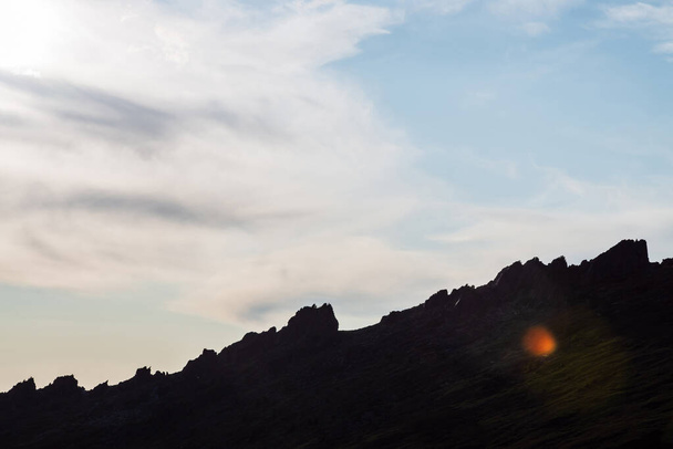 Sylwetka skał w podświetleniu nad pochmurnym niebem. Zielona góra ze skałami w słońcu z rozbłyskiem słonecznym. Malowniczy minimalistyczny krajobraz alpejski w słoneczny dzień. Piękne górskie krajobrazy z ciemnym wzgórzem. - Zdjęcie, obraz