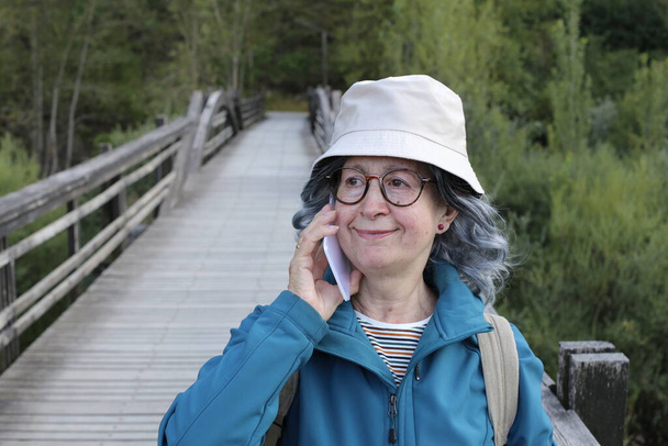 Nahaufnahme Porträt einer schönen reifen Frau, die ihr Smartphone im Freien auf einer Holzbrücke benutzt - Foto, Bild