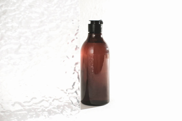 douchegel in een amberkleurige plastic fles naast transparante badkamerdeur. fles met waterdruppels in zonlicht. kopieer ruimte. hygiëne en dagelijks zelfzorgconcept - Foto, afbeelding