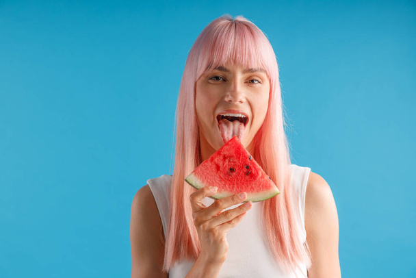 hravá mladá žena s růžovými vlasy drží plátek melounu, pózování s otevřenými ústy nad modrým studiem pozadí - Fotografie, Obrázek