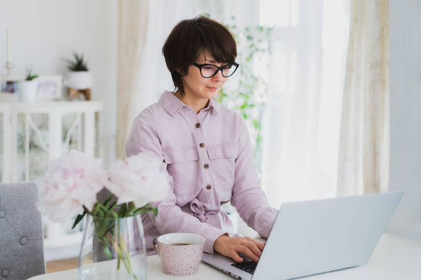Mixed race woman working at home using a laptop. Cozy workspace with fresh pink peonies flowers and a cup of tea on the white table with light interior background. Remote work at home. Copy space - Photo, image
