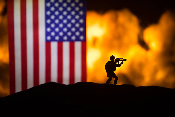 US small flag on burning dark background. Concept of crisis of war and political conflicts between nations. Silhouette of armed soldier against a USA flag. Selective focus - Photo, Image
