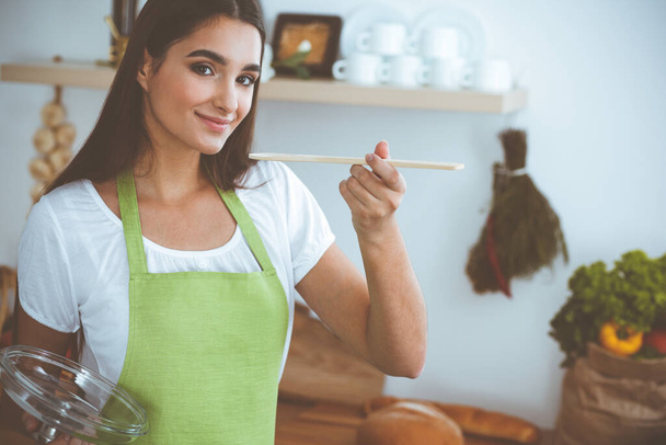 An attractive young dark-haired woman tasting ready hot meal with a wooden spoon while standing and smiling near the kitchen stove. Cooking and householding concepts. - Foto, afbeelding
