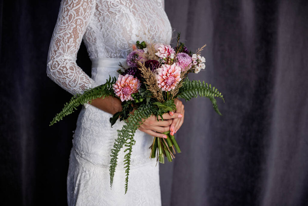 Gentle hands of the bride hold wedding bouquet of flowers against background of black fabric. The girl made bouquet of green ferns, pink chrysanthemums, dry pampas grass, purple Dahlias, white Alyssum - Fotó, kép
