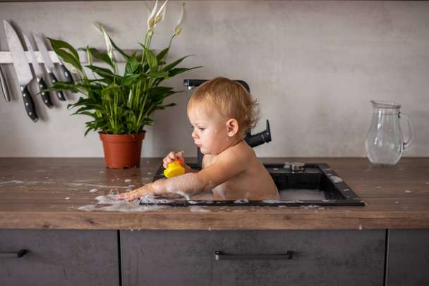 Χαριτωμένο χαρούμενο κοριτσάκι με το παιχνίδι με νερό και αφρό σε ένα νεροχύτη κουζίνα στο σπίτι - Φωτογραφία, εικόνα