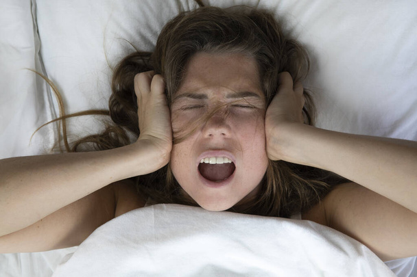 Mujer joven deprimida infeliz acostada en la cama con problemas de salud, gritando, resaca, depresión, mujer insomne, migraña vista superior con sábanas blancas de cerca - Foto, imagen