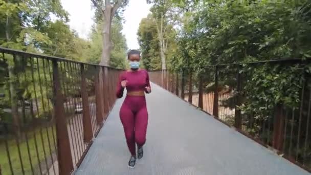 Mujer negra corriendo en puente con máscara médica protectora. Joven atlética hermosa hembra en ropa deportiva corriendo al aire libre durante el entrenamiento diario por la mañana. Coronavirus, concepto de contaminación atmosférica - Metraje, vídeo